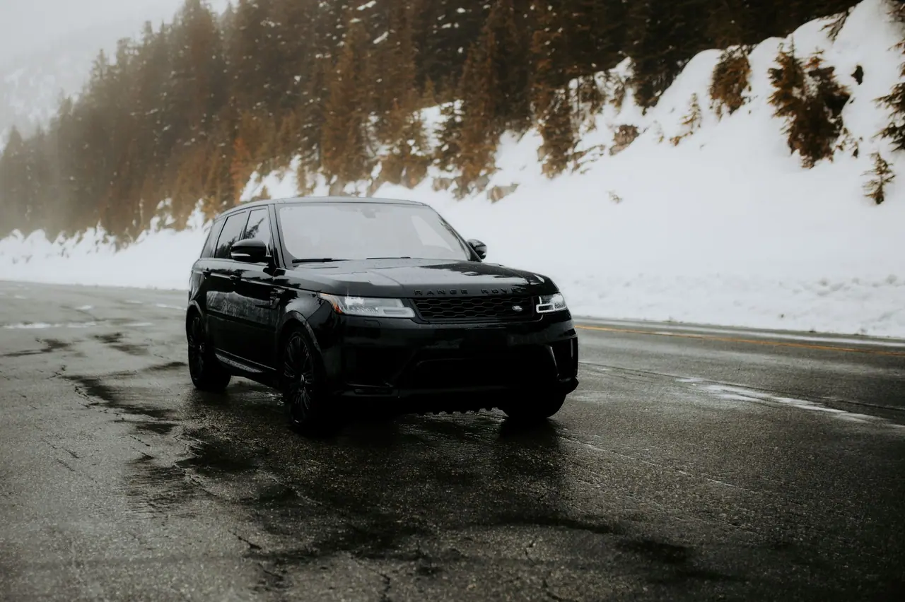 A sleek black SUV navigates a wet, snow-covered mountain road surrounded by evergreen trees.